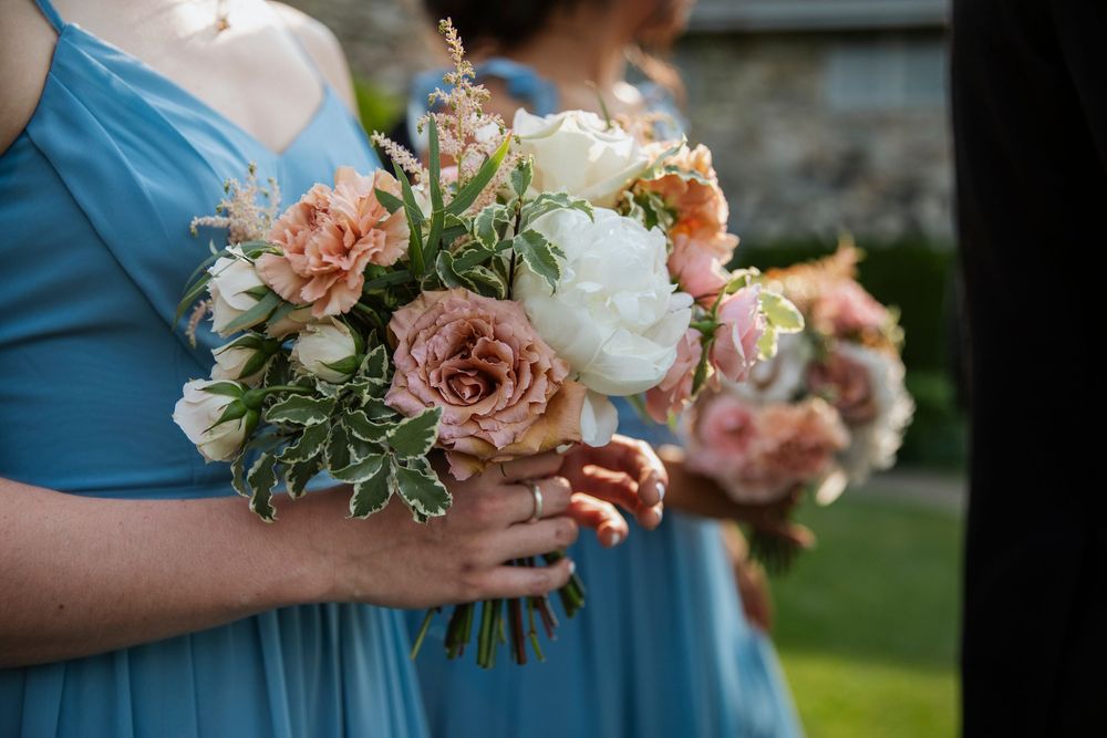 Bridesmaids in blue dresses hold elegant bouquets of pink roses, white peonies and greenery at an outdoor wedding.