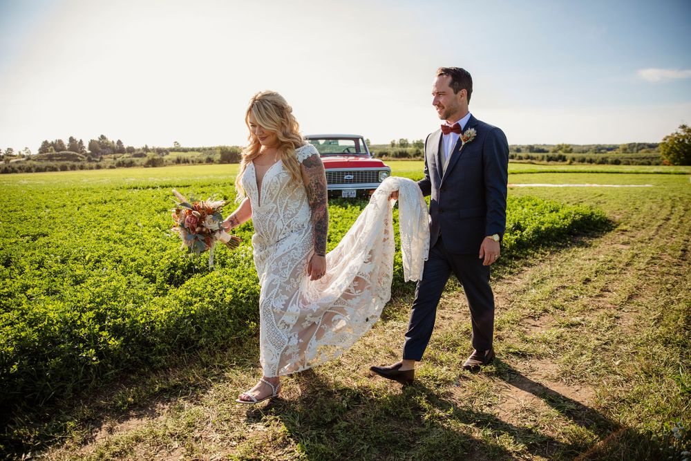 A couple in wedding attire walks together through a sunny agricultural field with golden light in the background.