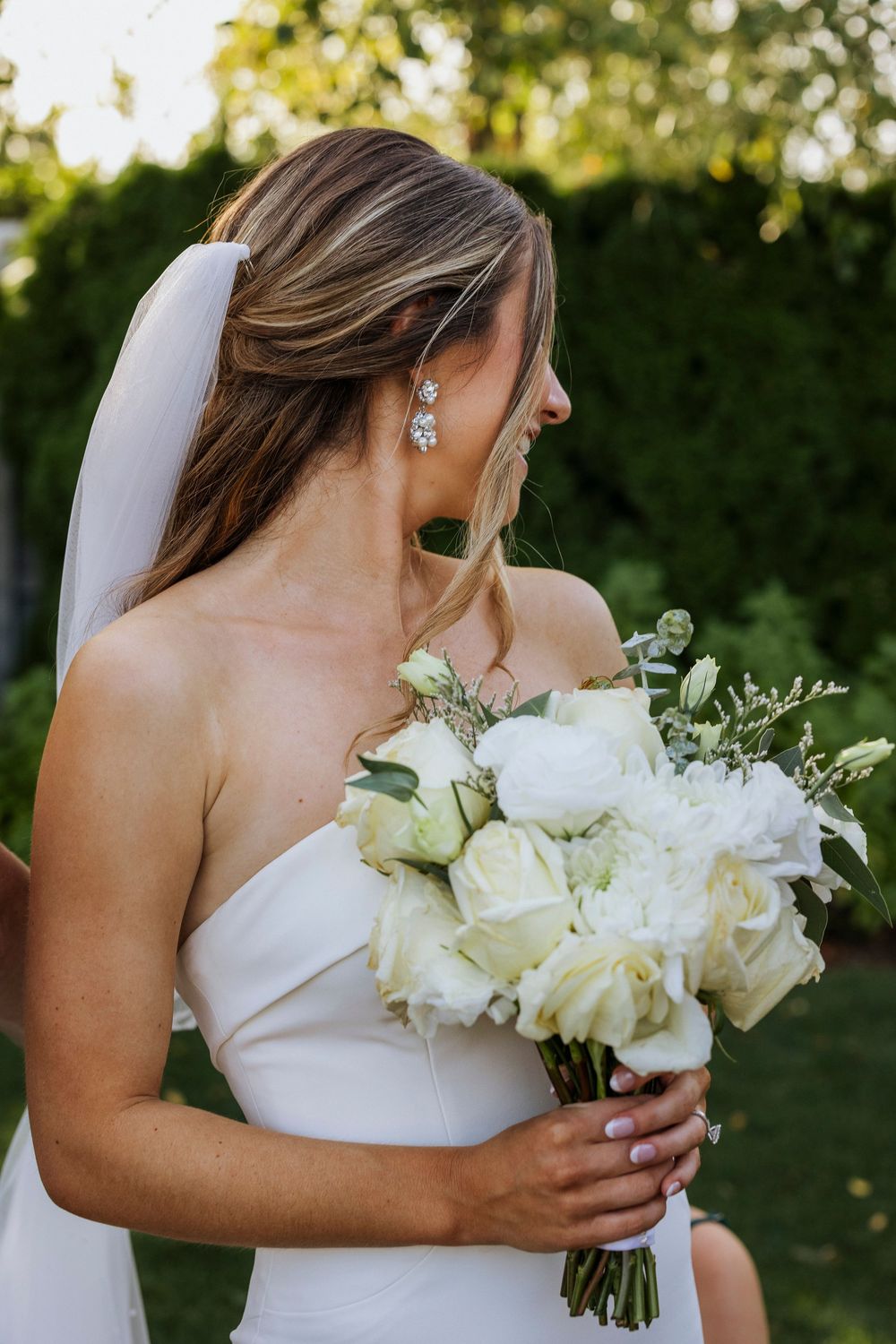 Bride in strapless white wedding dress holds elegant bouquet of white roses and greenery in outdoor garden setting.