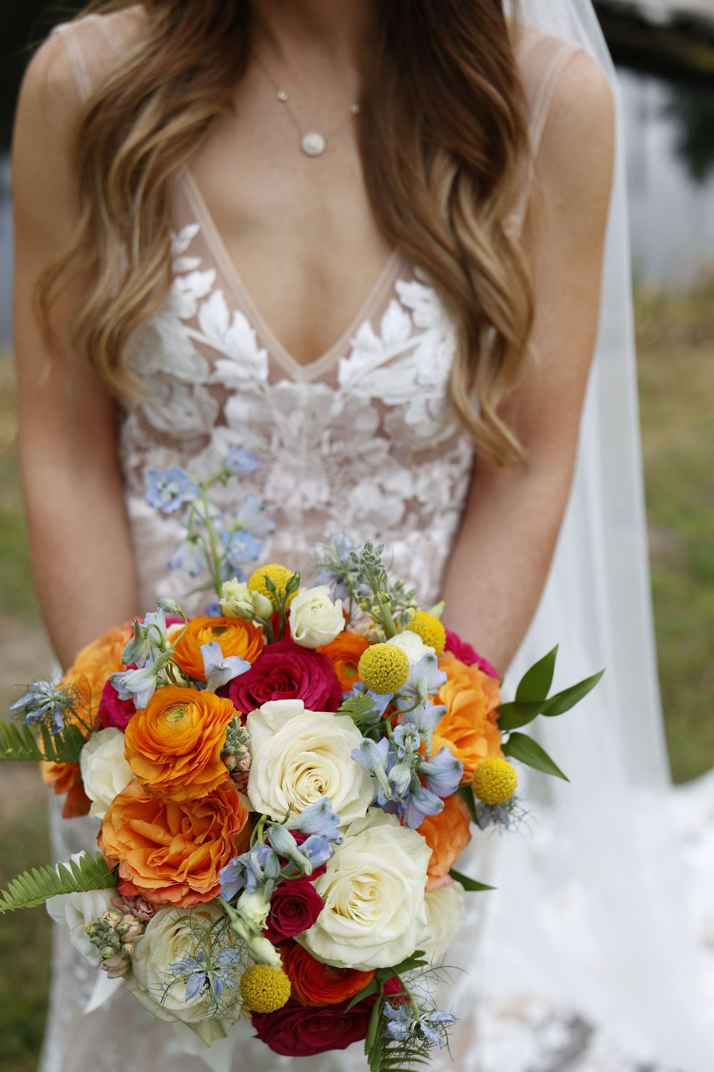 A vibrant wedding bouquet featuring orange ranunculus, white roses, and blue accents against a white bridal gown in Tawas City Michigan.