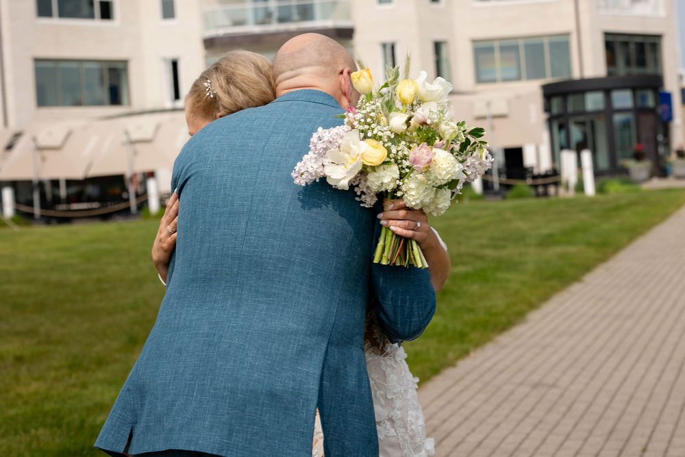 A dad in blue formal attire embrace while holding a bouquet of white and pink flowers on a grassy lawn at Bay Harbor Michigan.