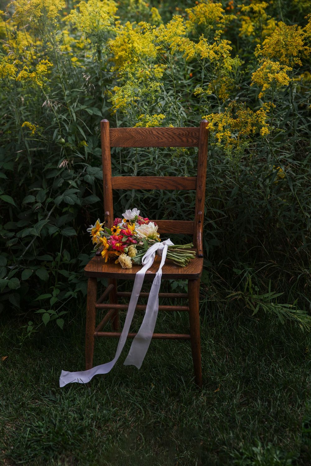 Rustic wooden chair adorned with colorful wildflower bouquet and white ribbons against a backdrop of golden flowers by Pachamama Floral.