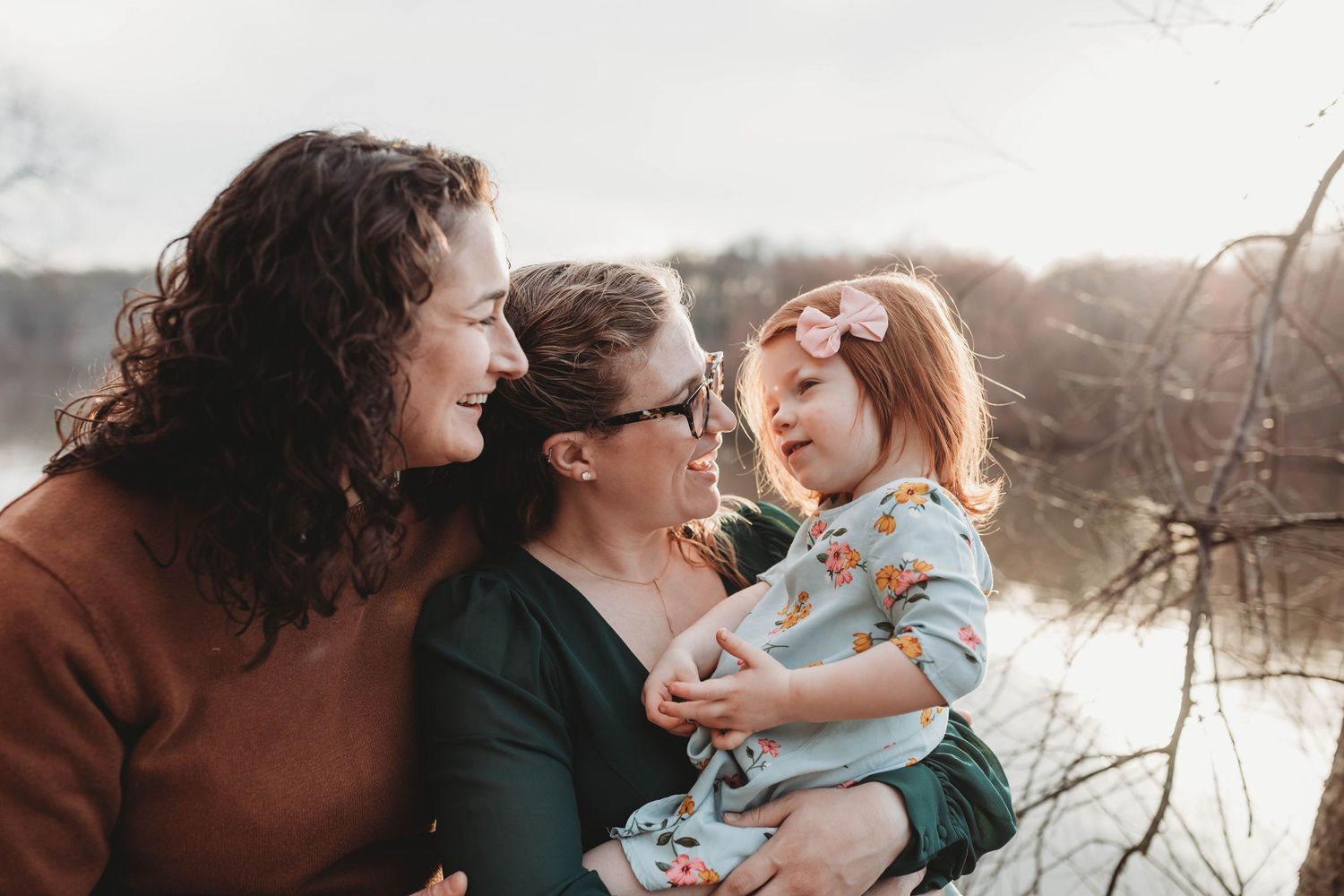 Two mothers smiling lovingly at their young red-haired daughter outdoors near a winter lake.