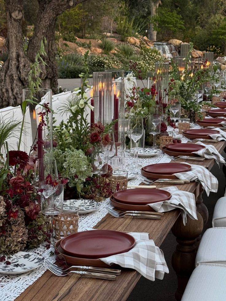 Rustic outdoor dining table set with burgundy plates, candles, and lush floral arrangements.