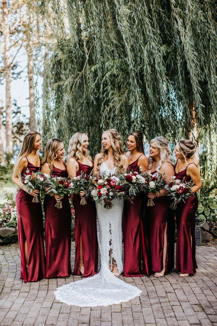 Bridesmaids in burgundy satin gowns holding lush floral bouquets stand with bride under weeping willow.
