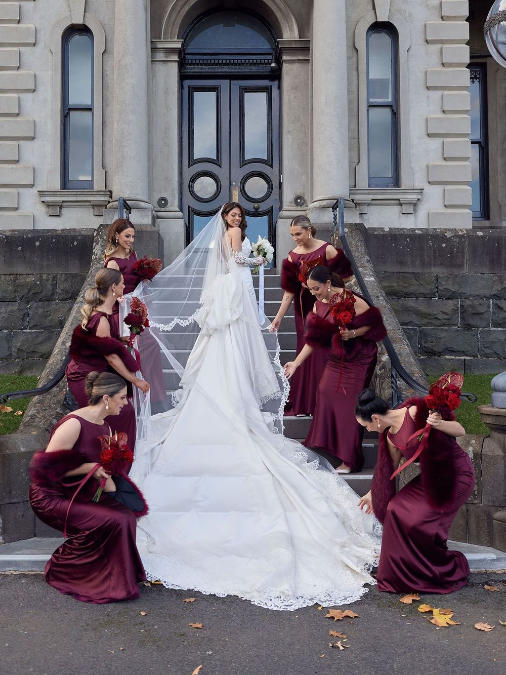 Bride in lace gown with long veil poses with bridesmaids in burgundy dresses on historic building steps.