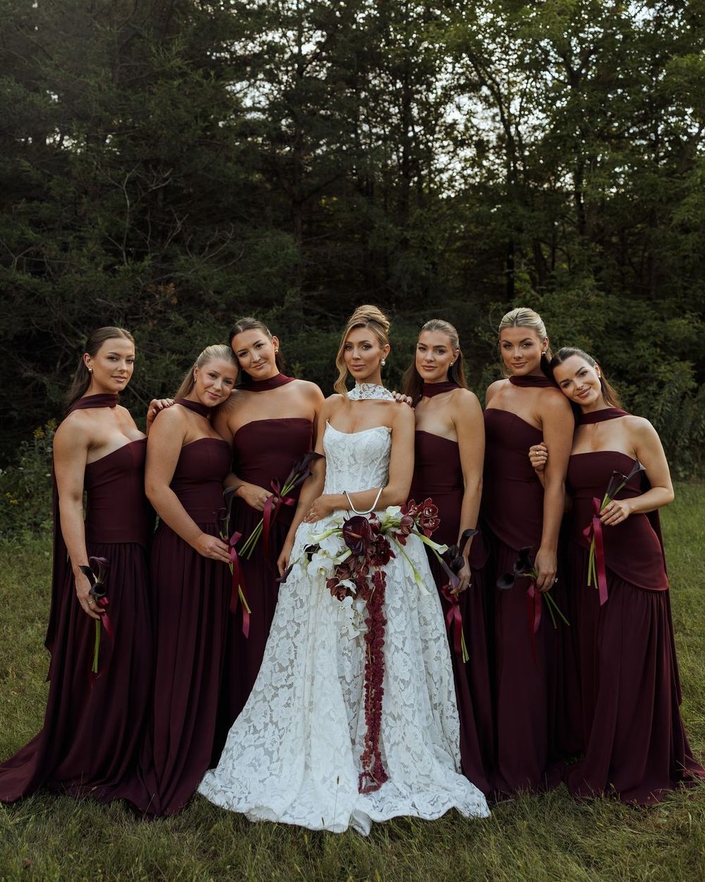 Bride in lace gown surrounded by bridesmaids in dark burgundy dresses holding cascading floral bouquet outdoors.