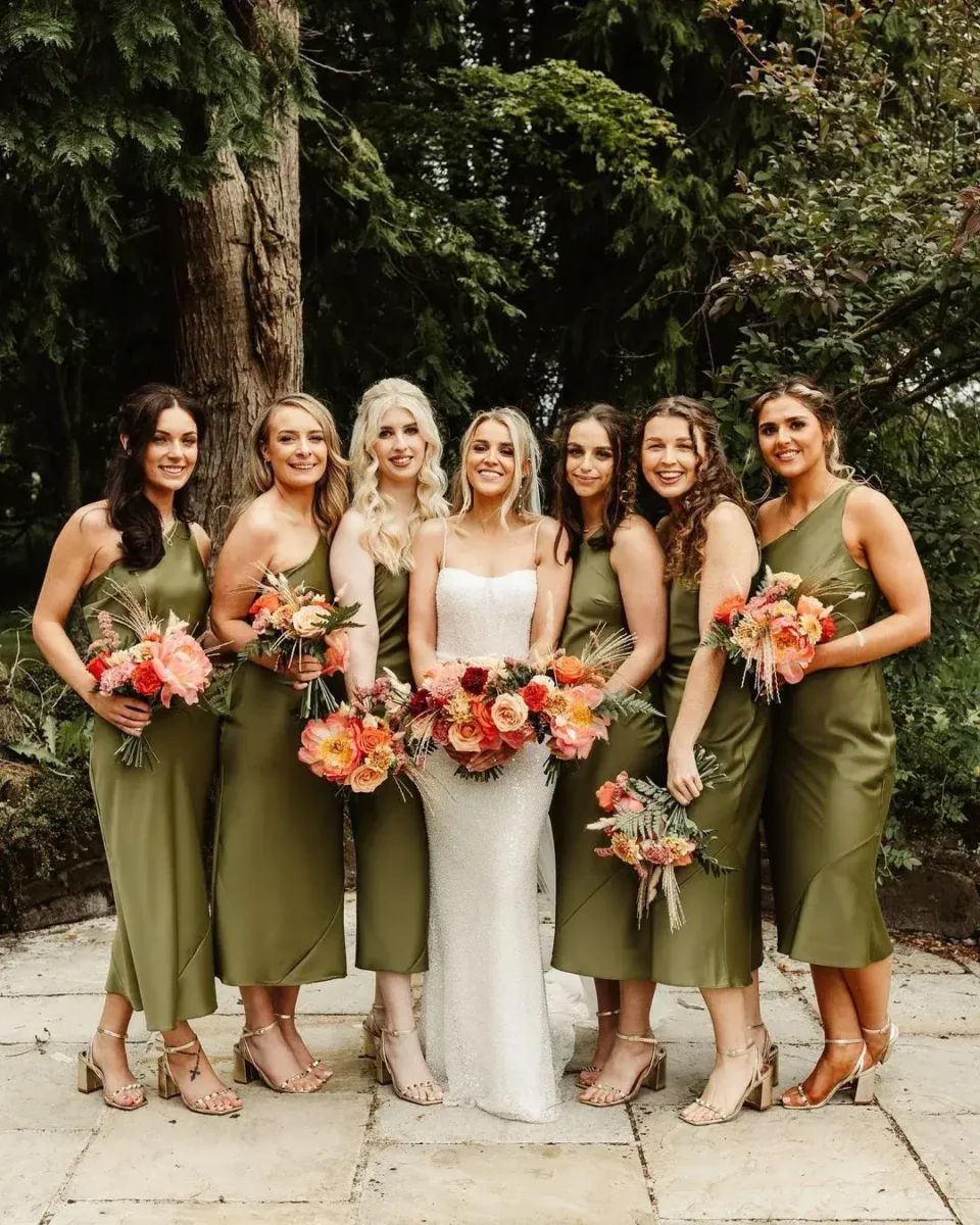 Bridesmaids in olive green midi satin dresses holding coral and orange bouquets stand with bride outdoors.