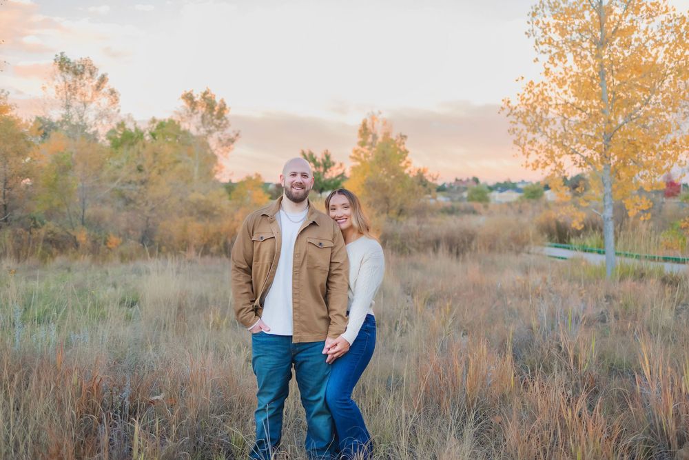 Couple walking through golden autumn field at sunset with tall grass and trees in the background.