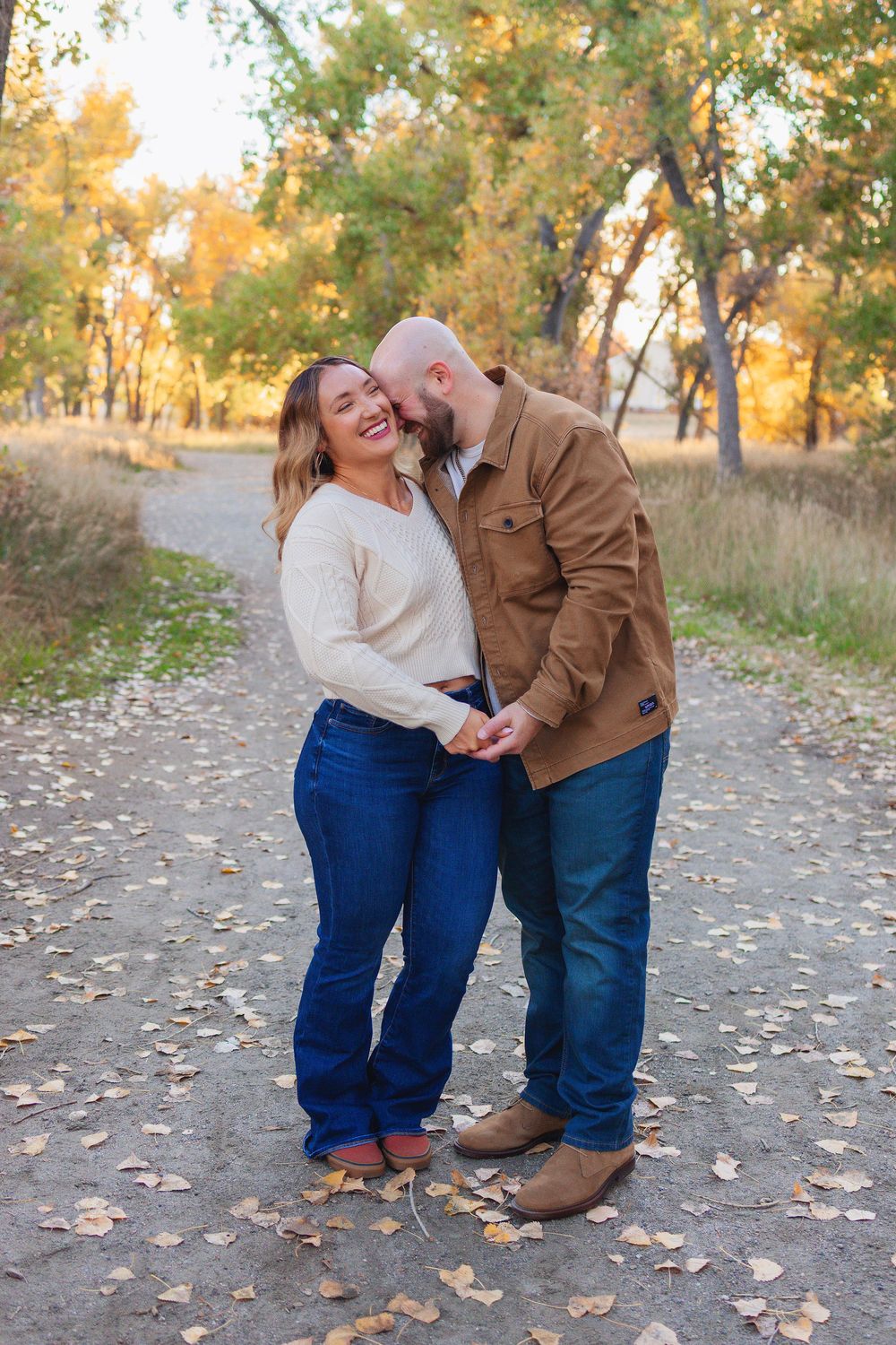 A couple shares a romantic moment on a path covered in autumn leaves with golden trees in the background.