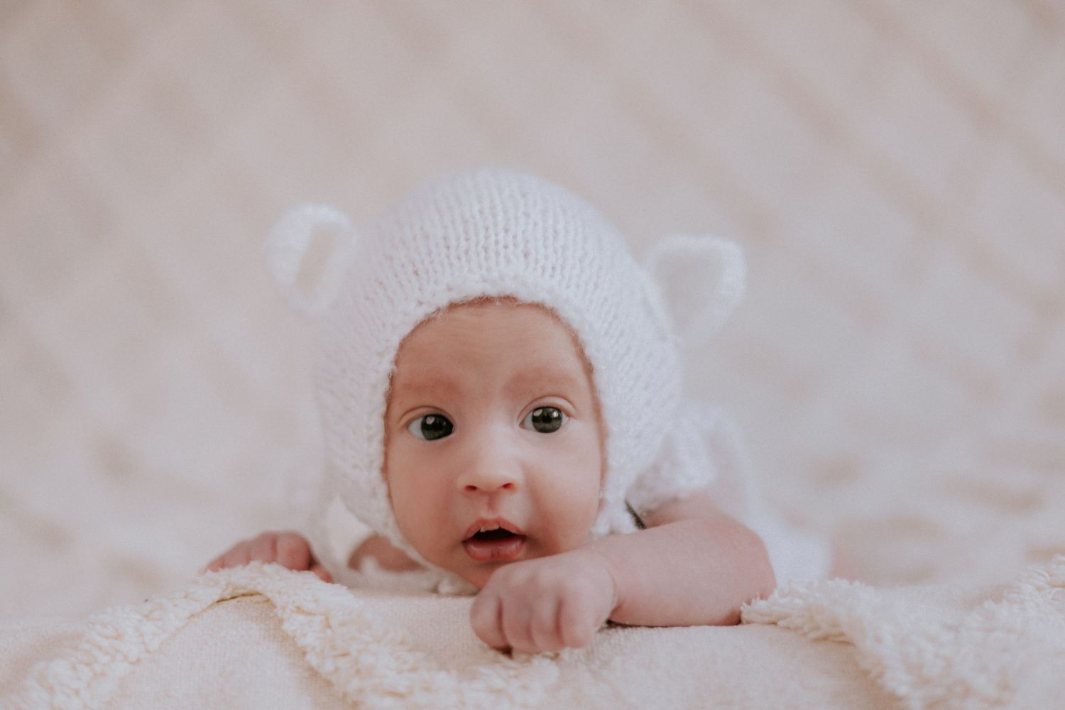 Adorabilné novorodenecké bábätko v bielej pletenej čiapočke s medvedíkovými ušami na jemnej krémovej deke. Rajecká dolina.