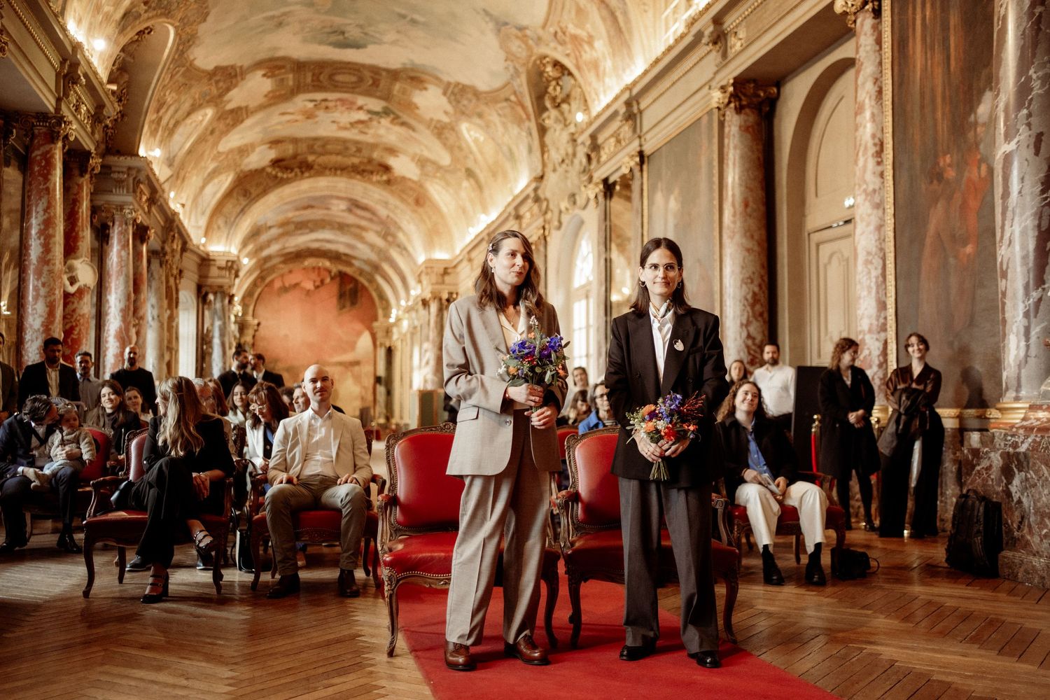 Guests mingle in an ornate baroque palace hallway with gilded decorations and red carpet during an elegant event.