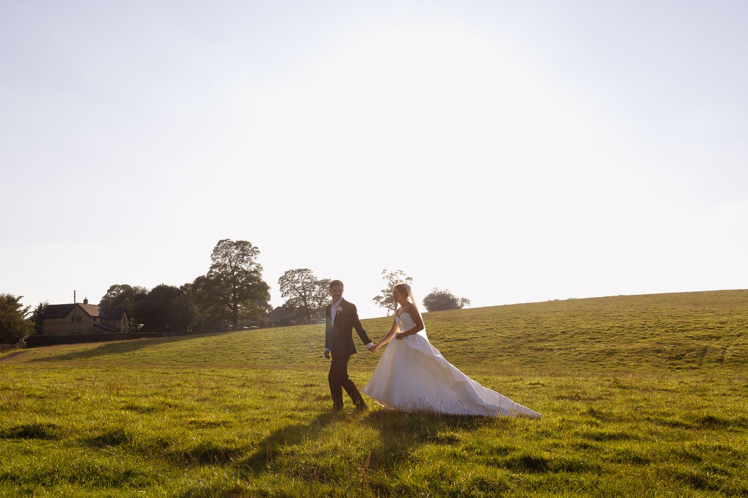 Couple in wedding attire walks across a sunlit grassy hill during golden hour.