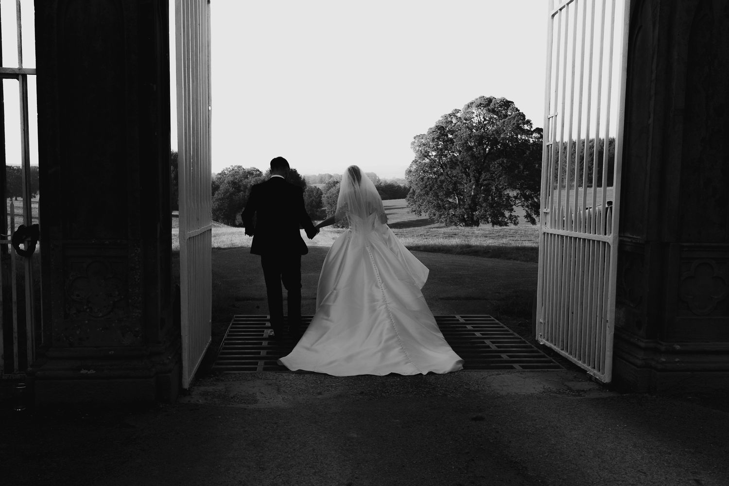 A bride in a flowing white wedding gown stands in silhouette at the end of a long hallway with columns.