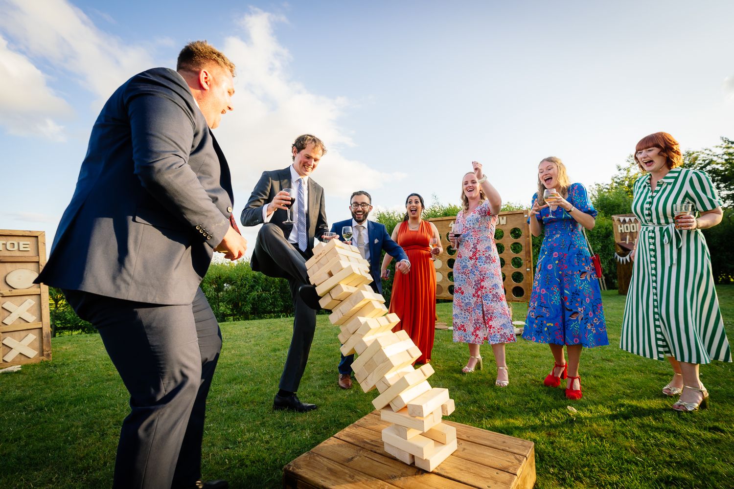 Outdoor wedding party watches as guests play giant wooden block game on a sunny lawn.