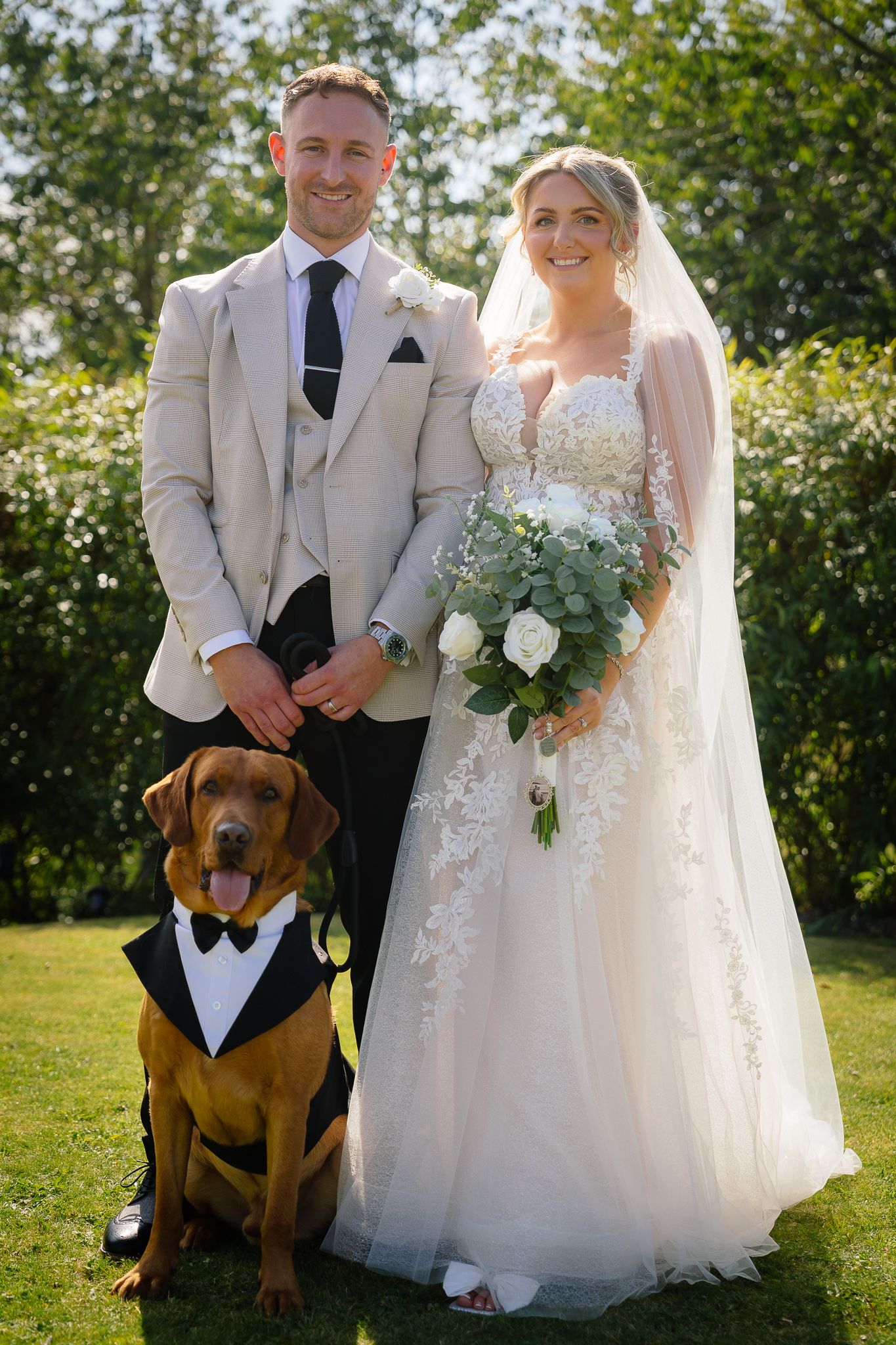 A brown dog wearing a formal bowtie poses with wedding couple in formal attire on sunny outdoor lawn.
