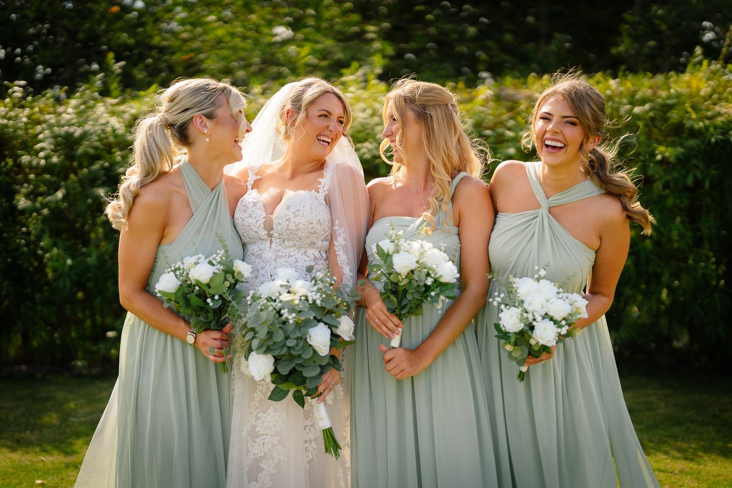 A wedding party wearing sage green bridesmaid dresses poses outdoors with white floral bouquets.
