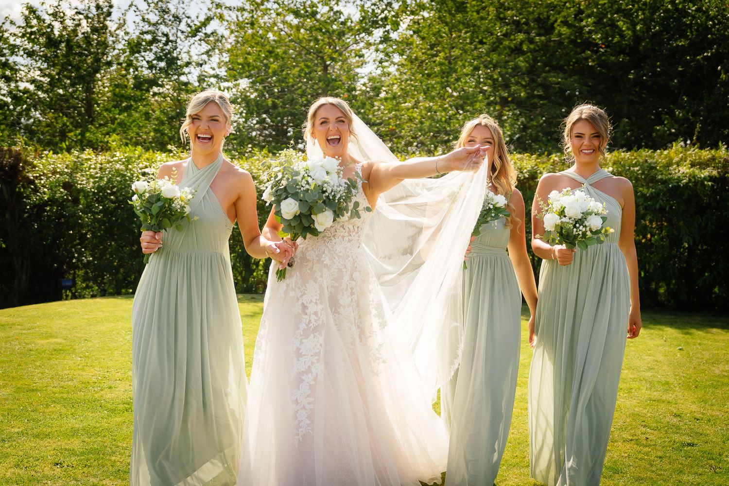 A bridal party in sage green bridesmaid dresses and white wedding gown walk together on a sunny garden lawn.