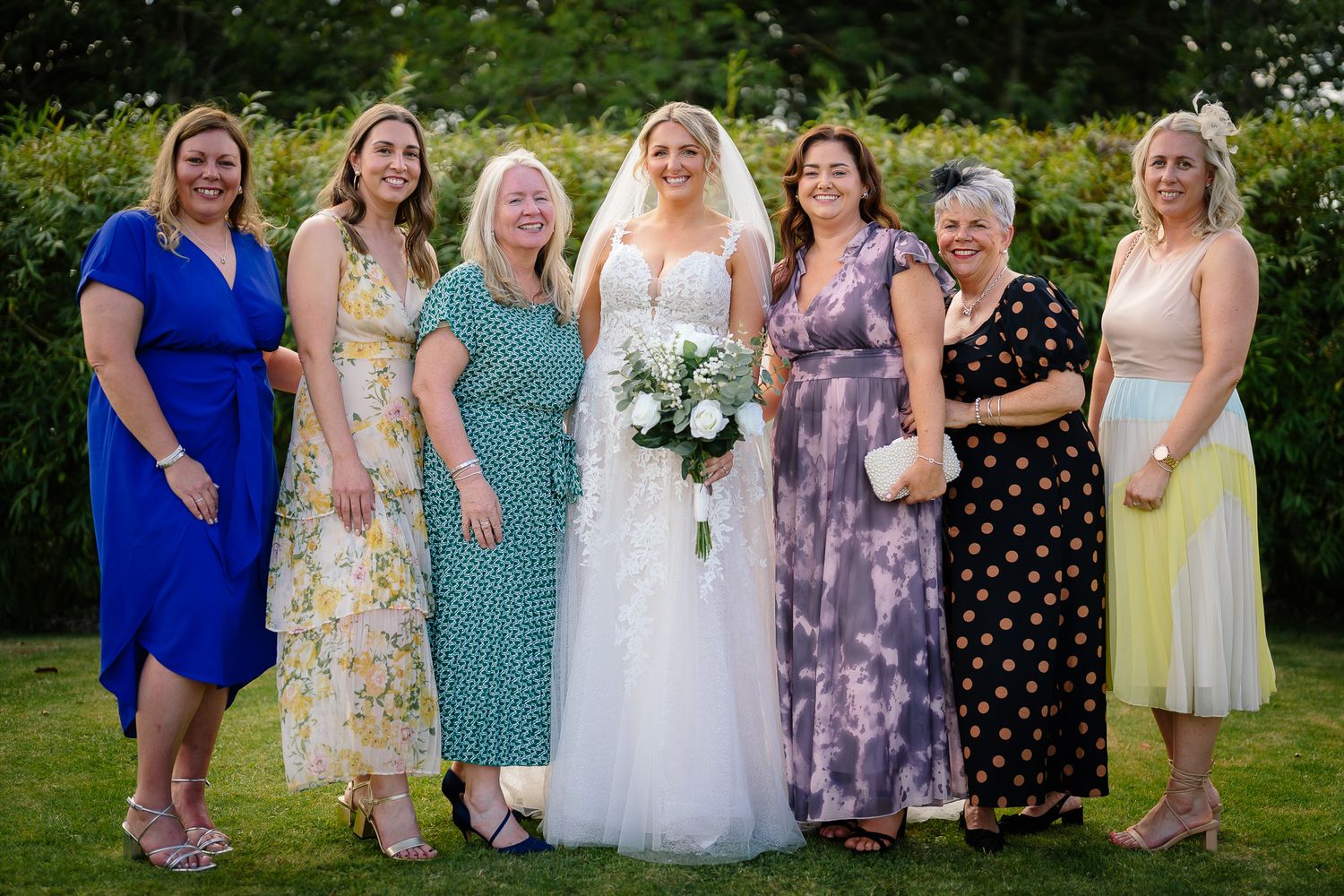 A group of people in formal attire gather outdoors for a wedding photo on a sunny summer day.