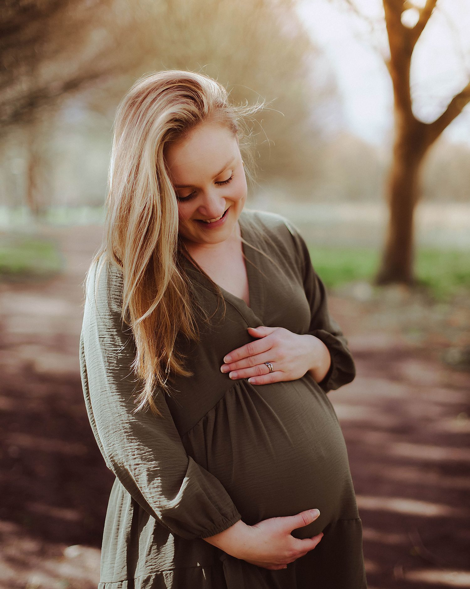 A pregnant women in a dark dress smiles while gently touching their baby bump in a sunlit outdoor setting. During a photoshoot in Bexley