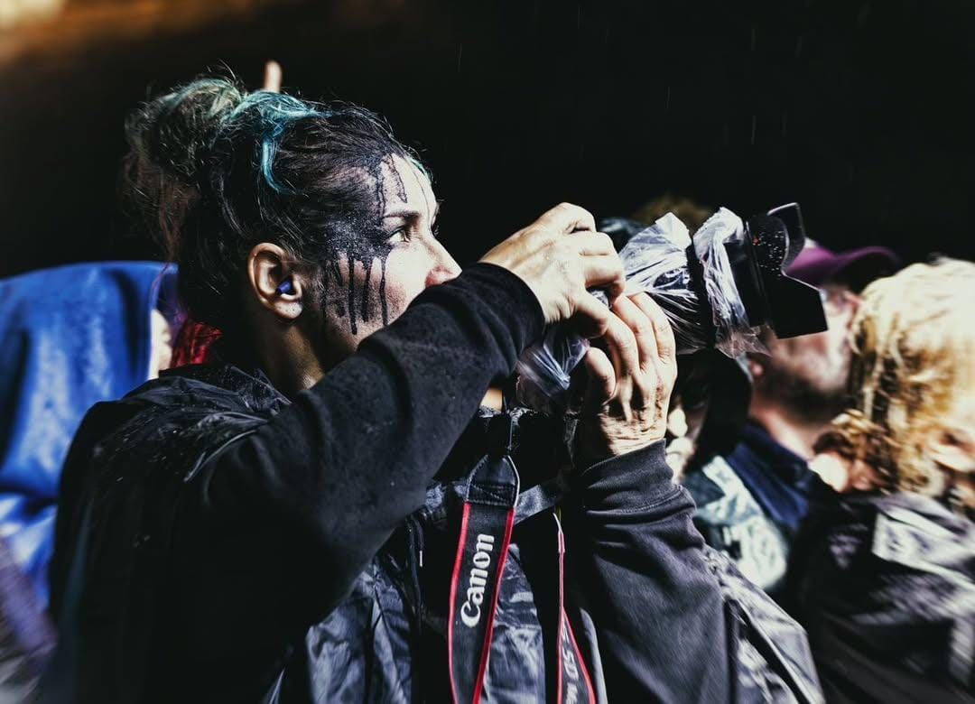 A photographer wearing dark clothing holds up a professional camera with a lens taking a shot in low light conditions.