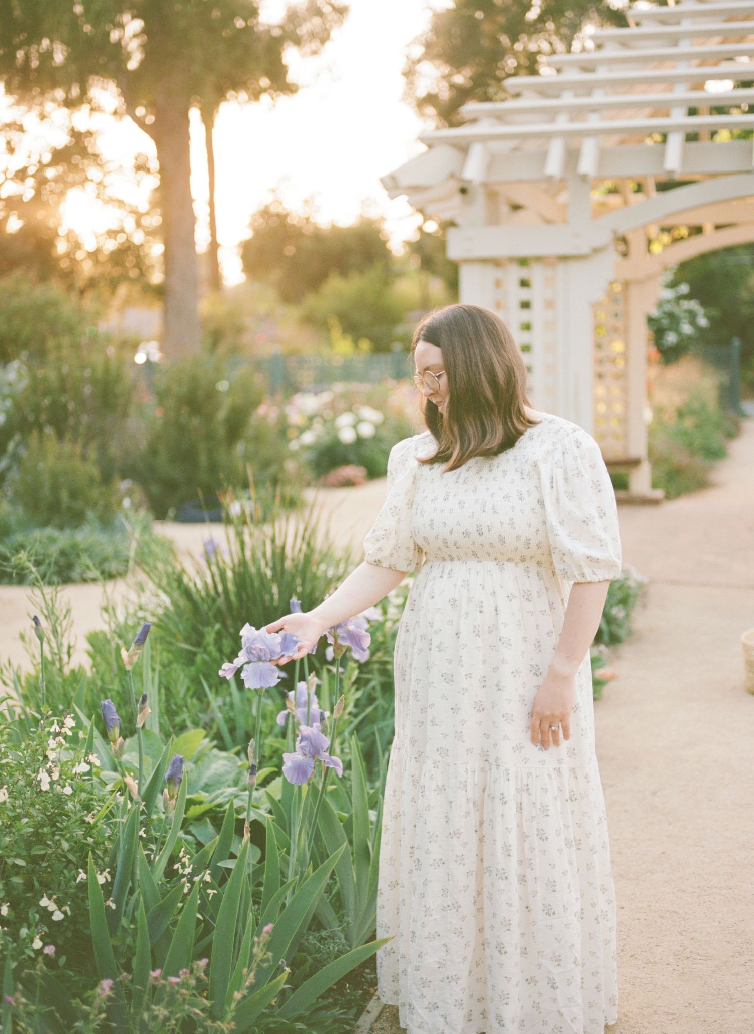 A person in a flowing white dress walks along a garden path at sunset among blooming purple irises.