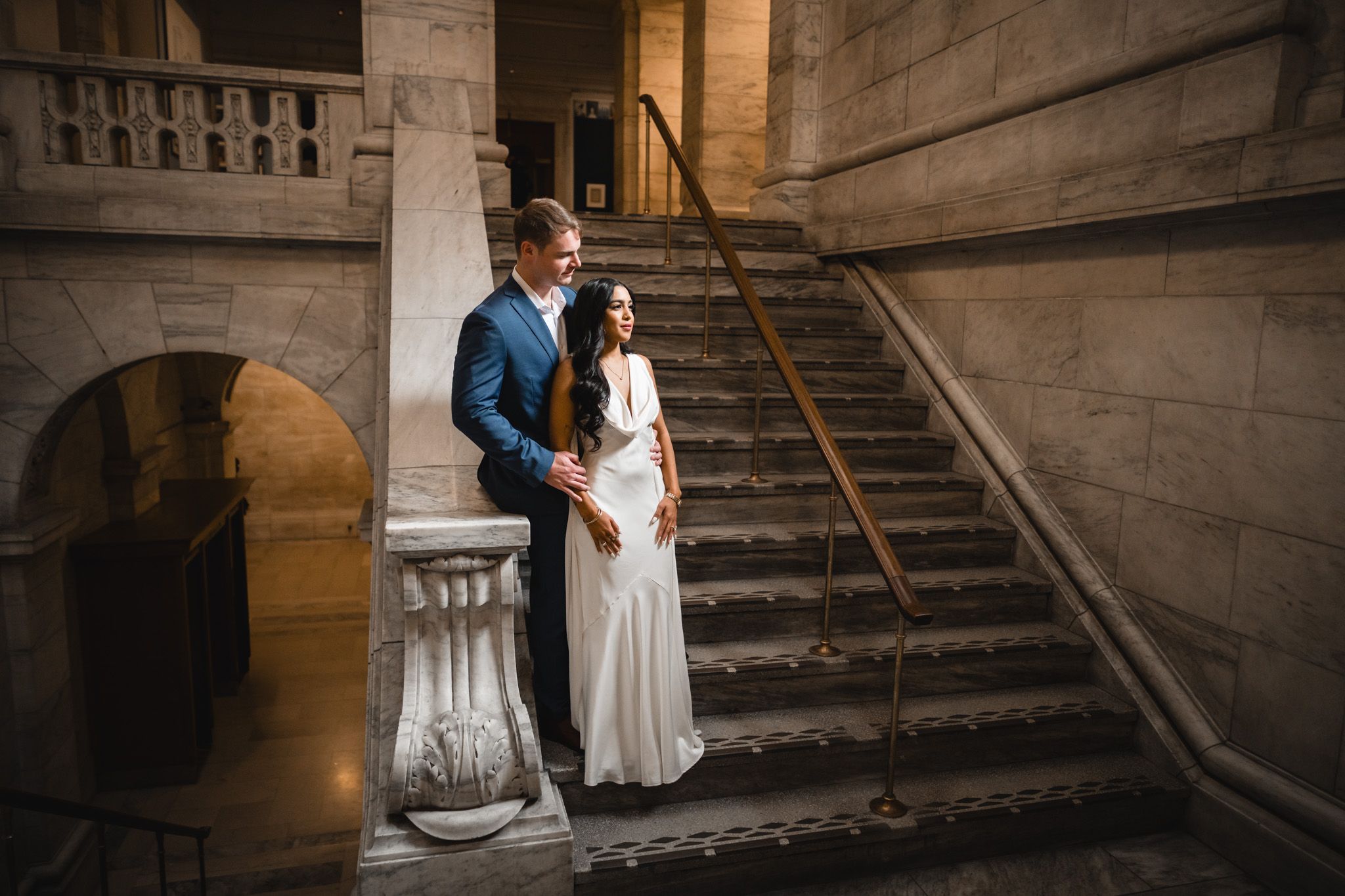 Elegant couple poses on grand marble staircase in romantic wedding photoshoot.