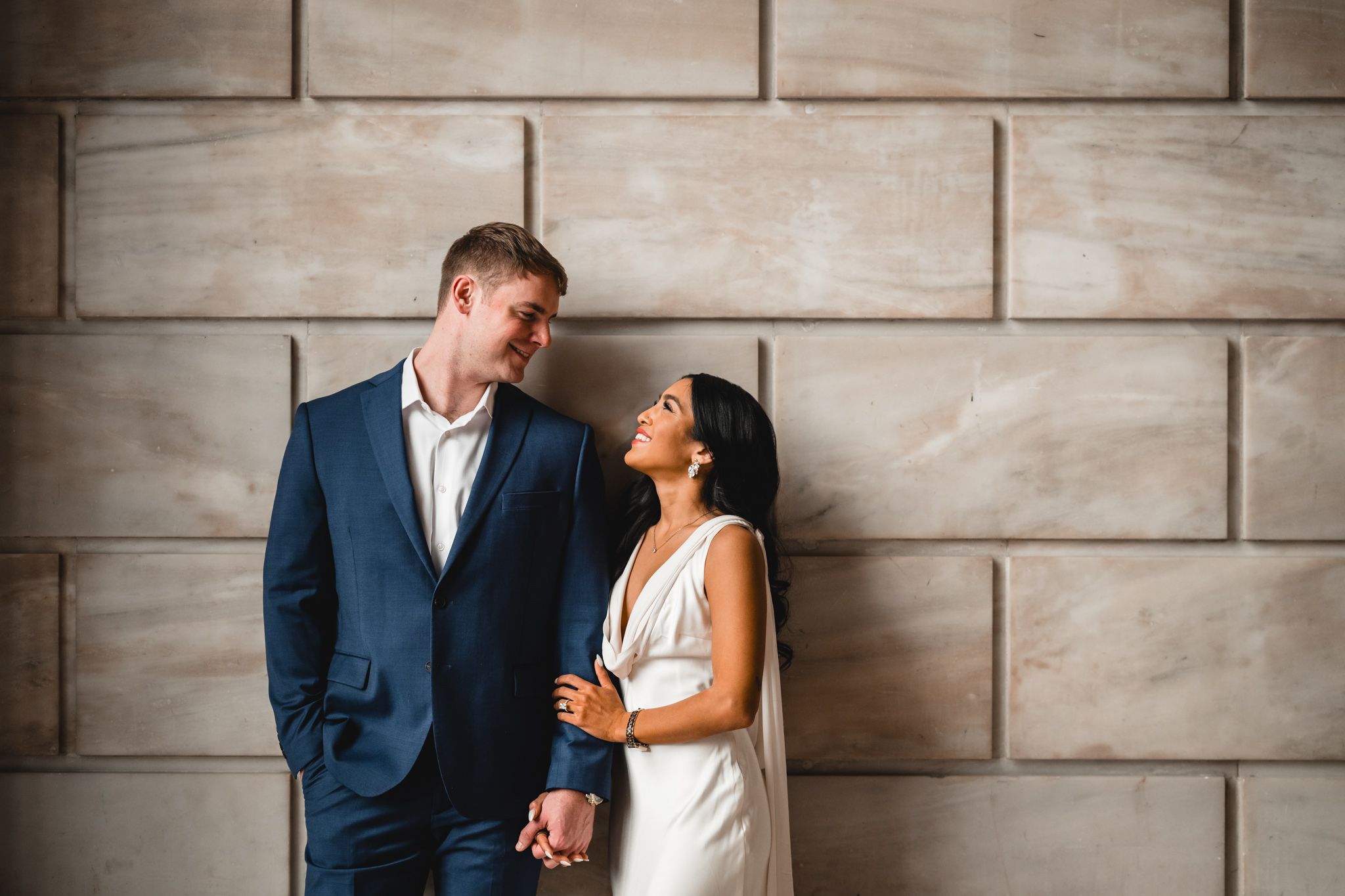 Couple in formal attire embraces while posing against a textured stone wall.