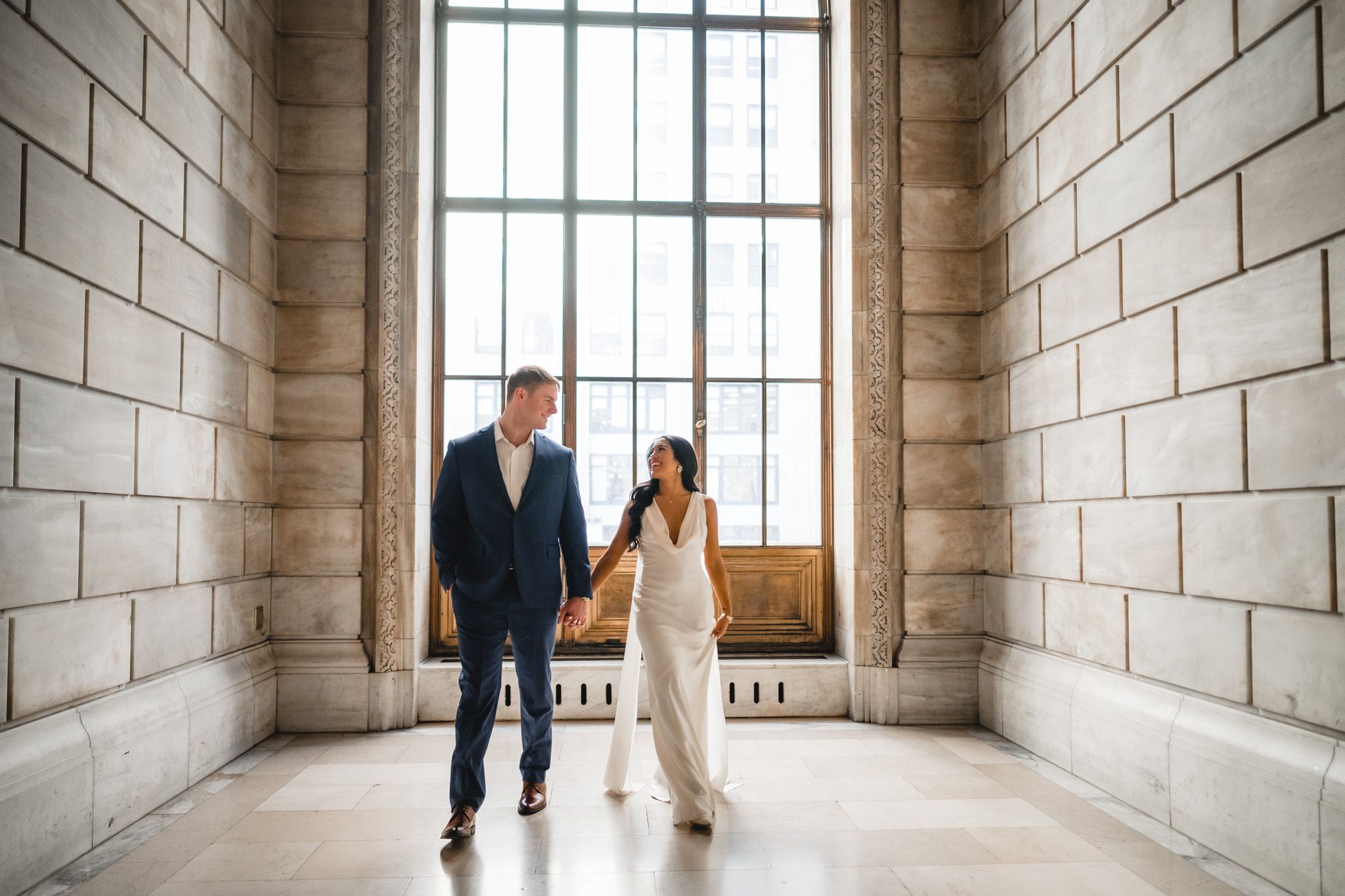 Couple walks together in an elegant stone hallway with large windows.