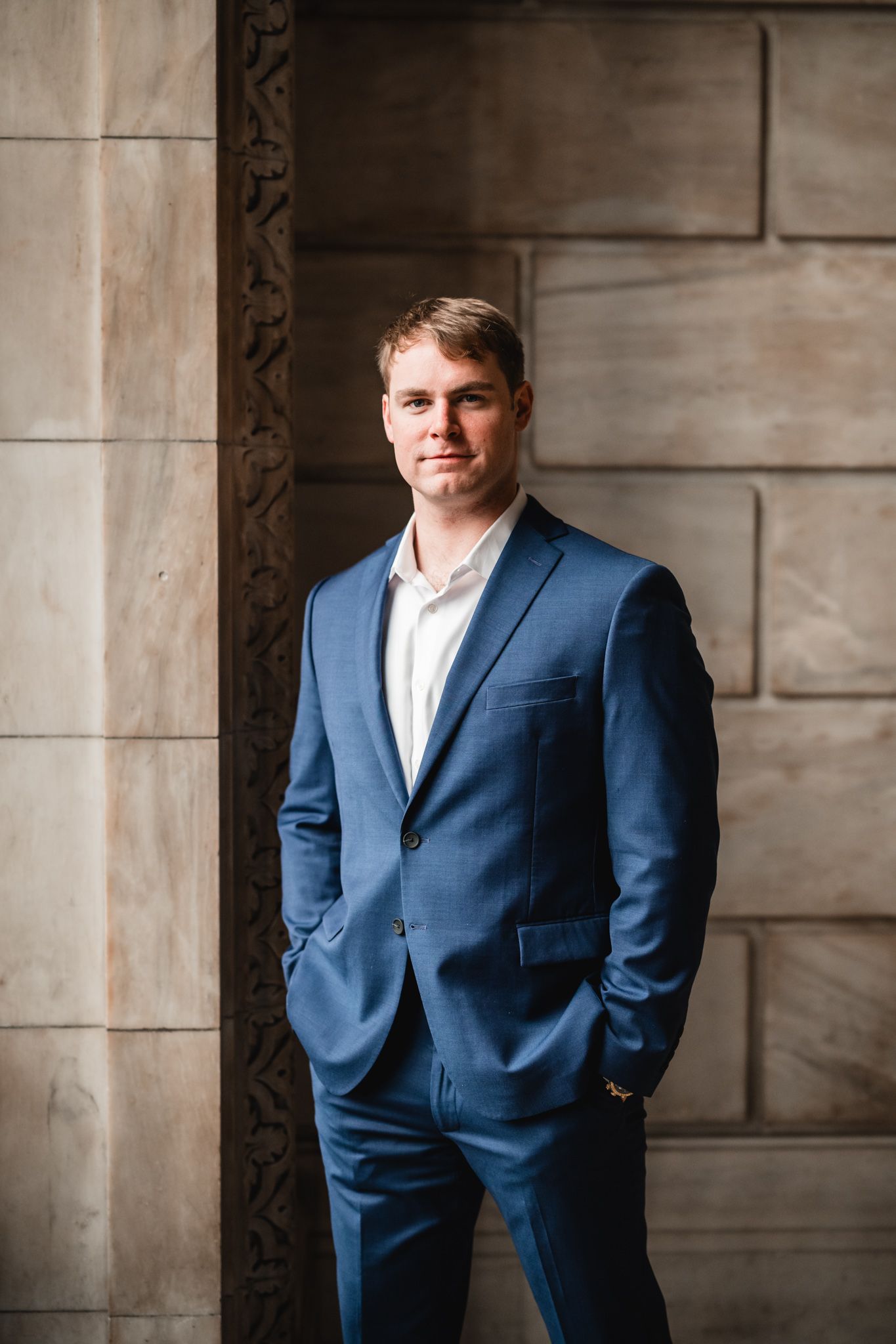 Person in navy suit poses confidently against stone wall backdrop.