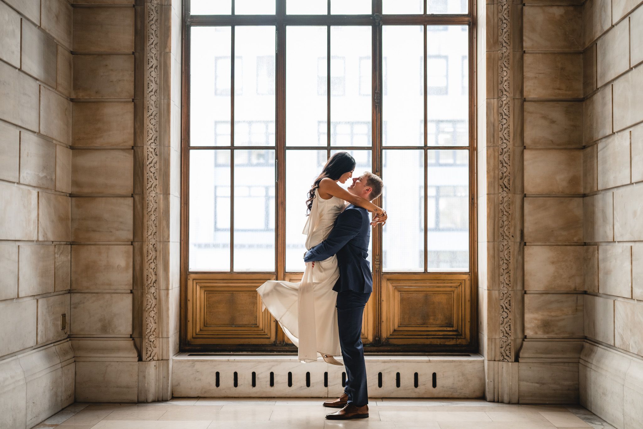 Couple silhouetted against bright windows in historic stone corridor.