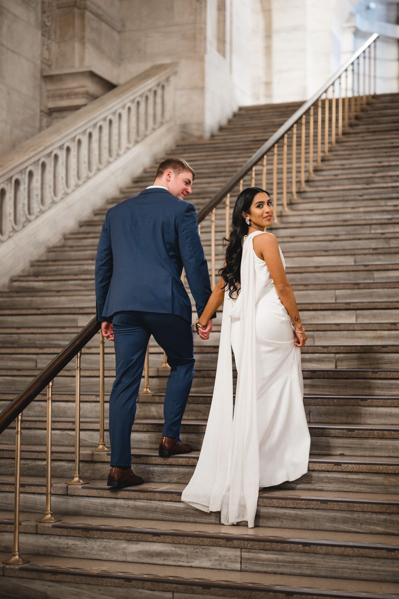 Couple in formal attire ascends grand stone staircase in historic building.