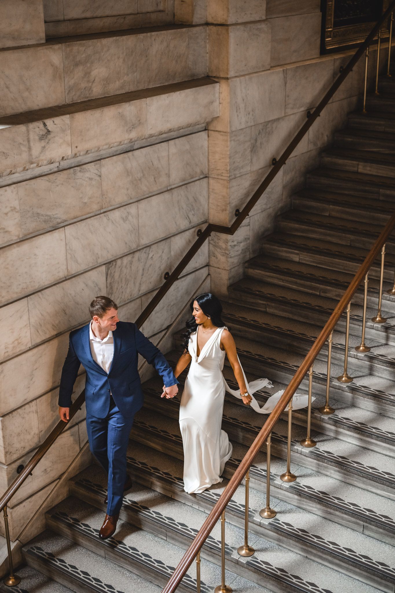 Couple descends elegant marble staircase in classic wedding attire.