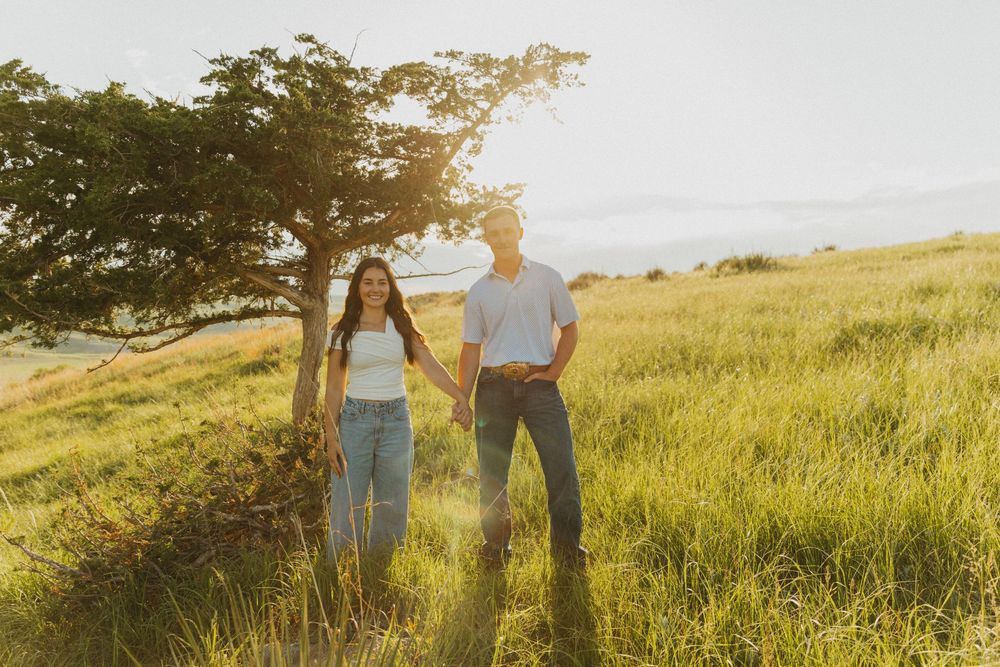 A couple stands in a golden sunlit meadow at sunset holding hands while wearing casual attire.