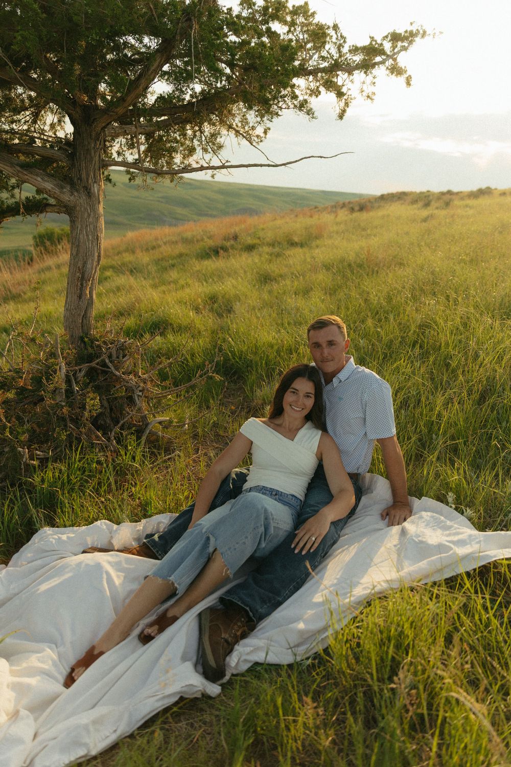 A couple relaxes on a white blanket in a scenic hilltop meadow during golden hour.