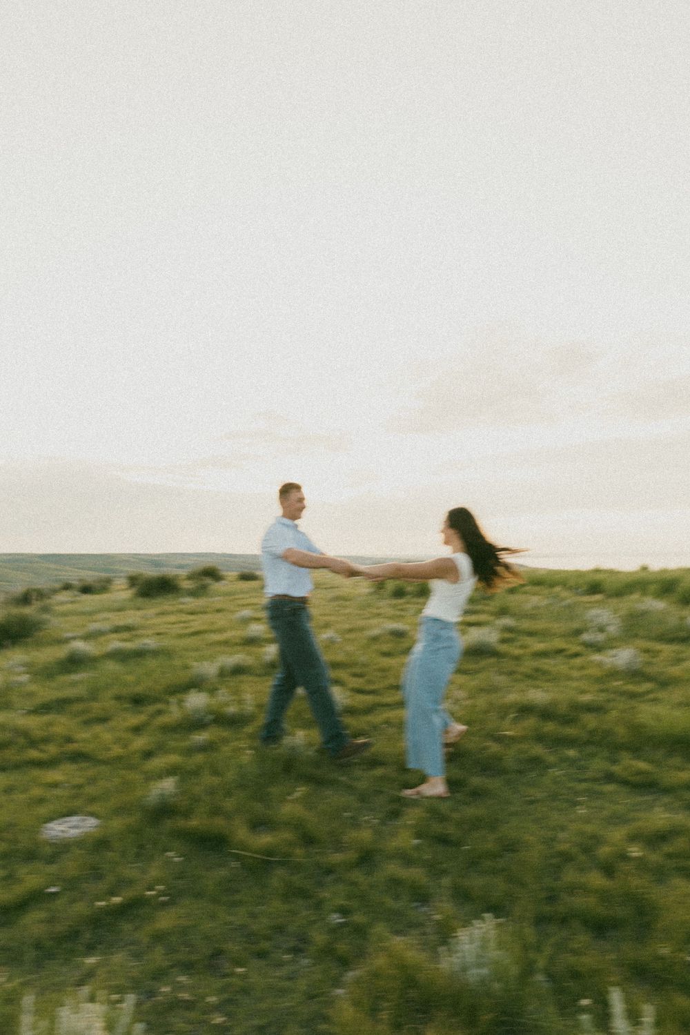A couple spins and dances together on a grassy hilltop at sunset in a playful moment.