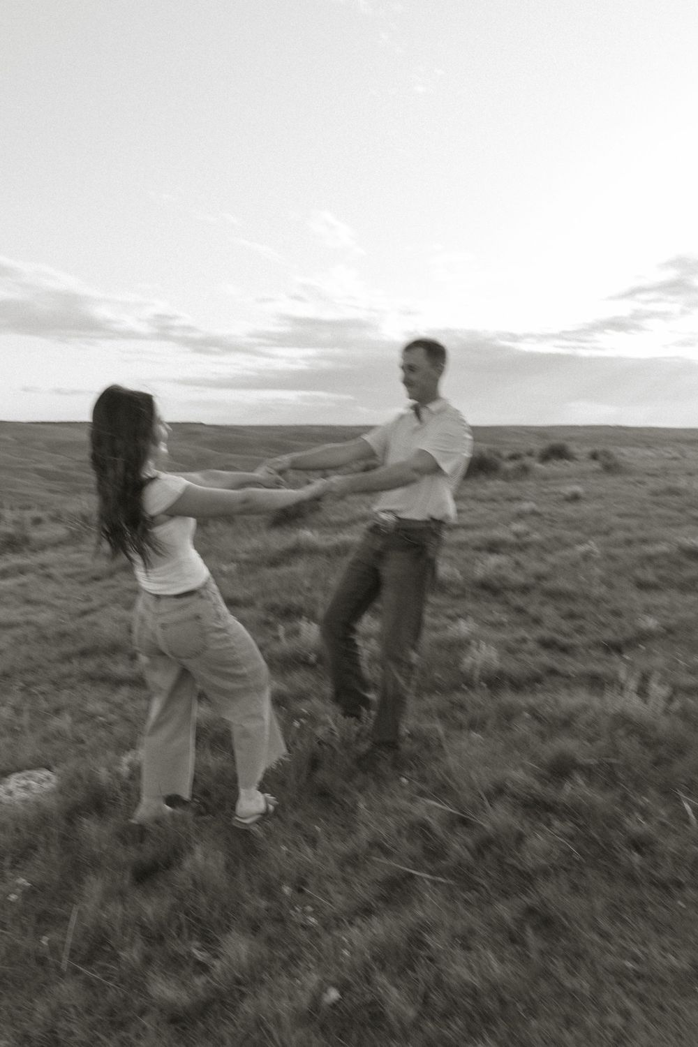 A couple dances together in a grassy field at sunset, captured in black and white photography.