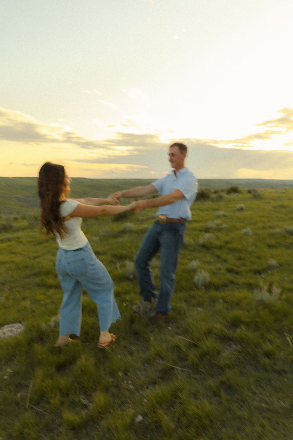 A couple dances playfully together in a grassy field at sunset with rolling hills in the background.
