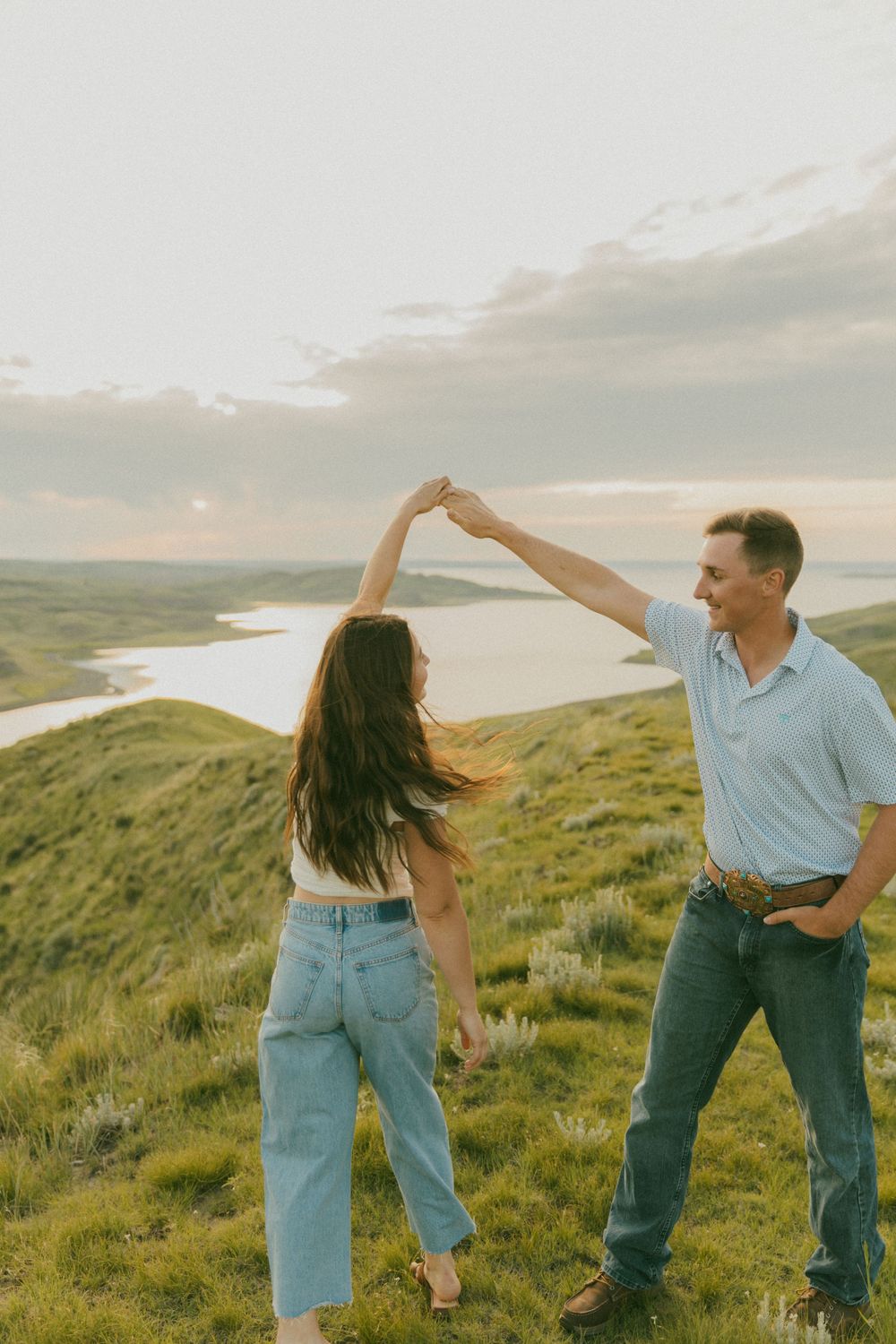 Playful couple dances and poses together on a grassy hillside overlooking a lake.