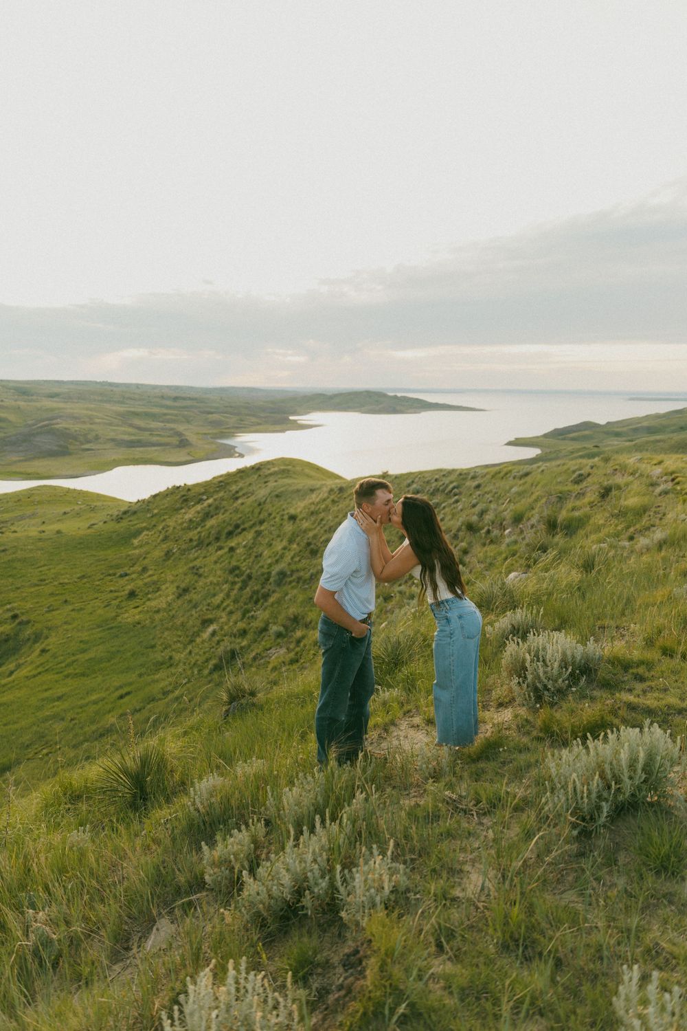 Two people stand on a scenic hillside overlooking a serene lake surrounded by rolling green hills at sunset.