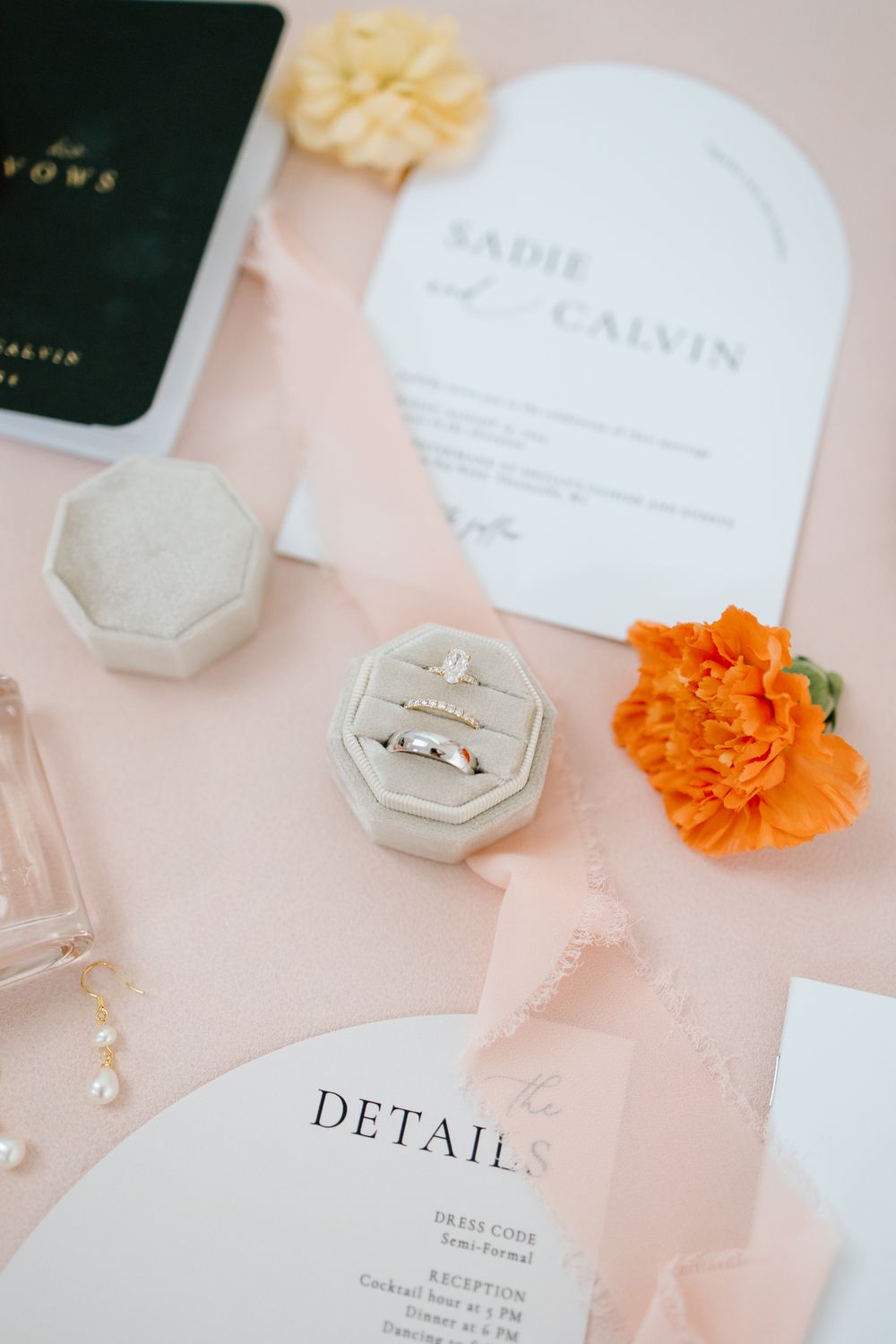 Wedding invitation details with orange marigold flowers and velvet ring box on a soft pink background.