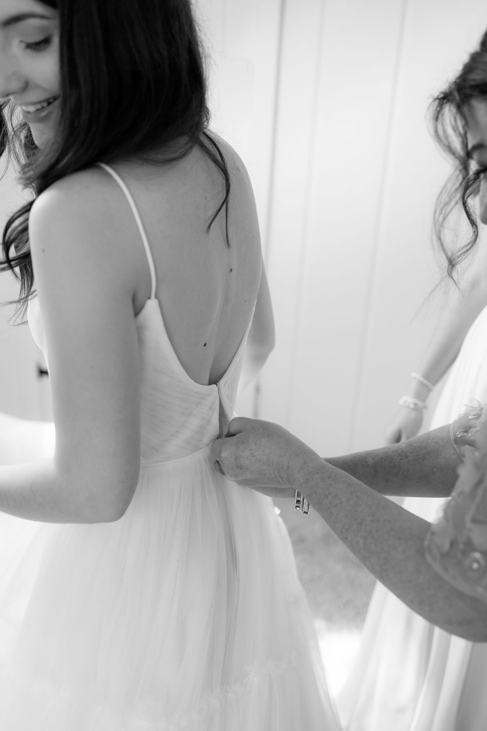 A bride in a white dress gets ready on her wedding day while surrounded by bridesmaids in a bright room.