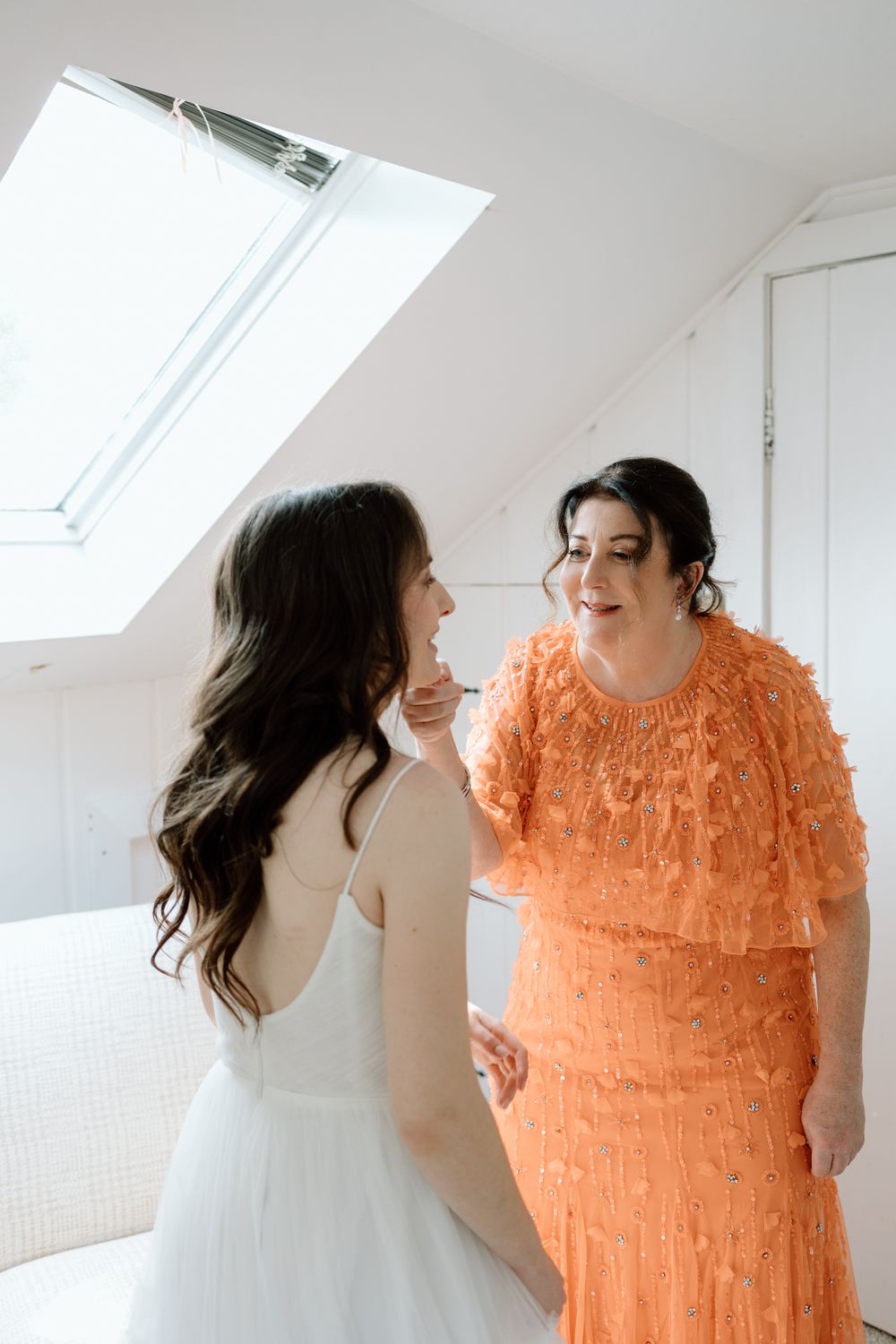 Two friends in white and orange dresses laugh together in a bright room with skylights.