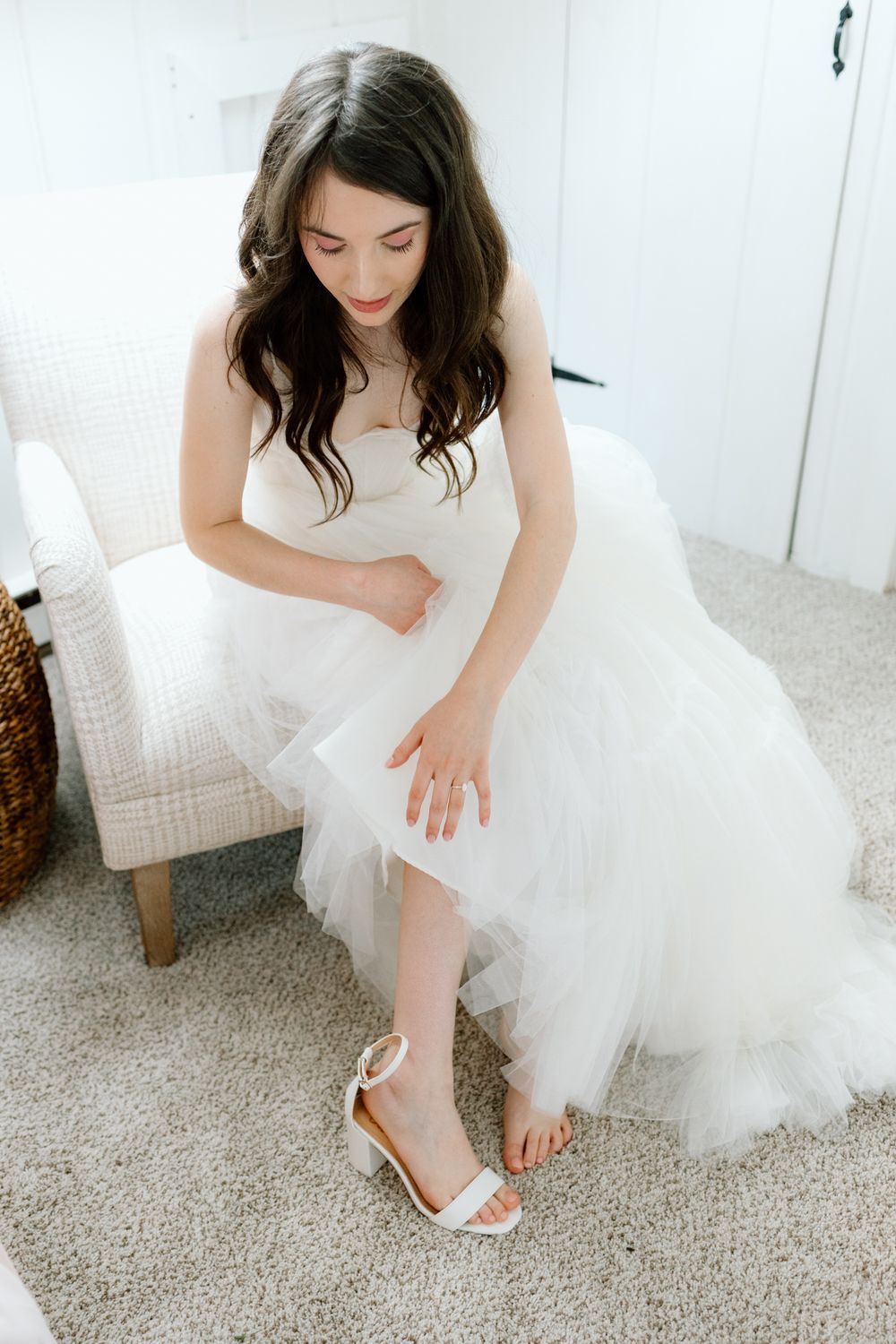 Bride in flowing white wedding dress adjusts delicate strappy sandals while sitting on carpeted floor.