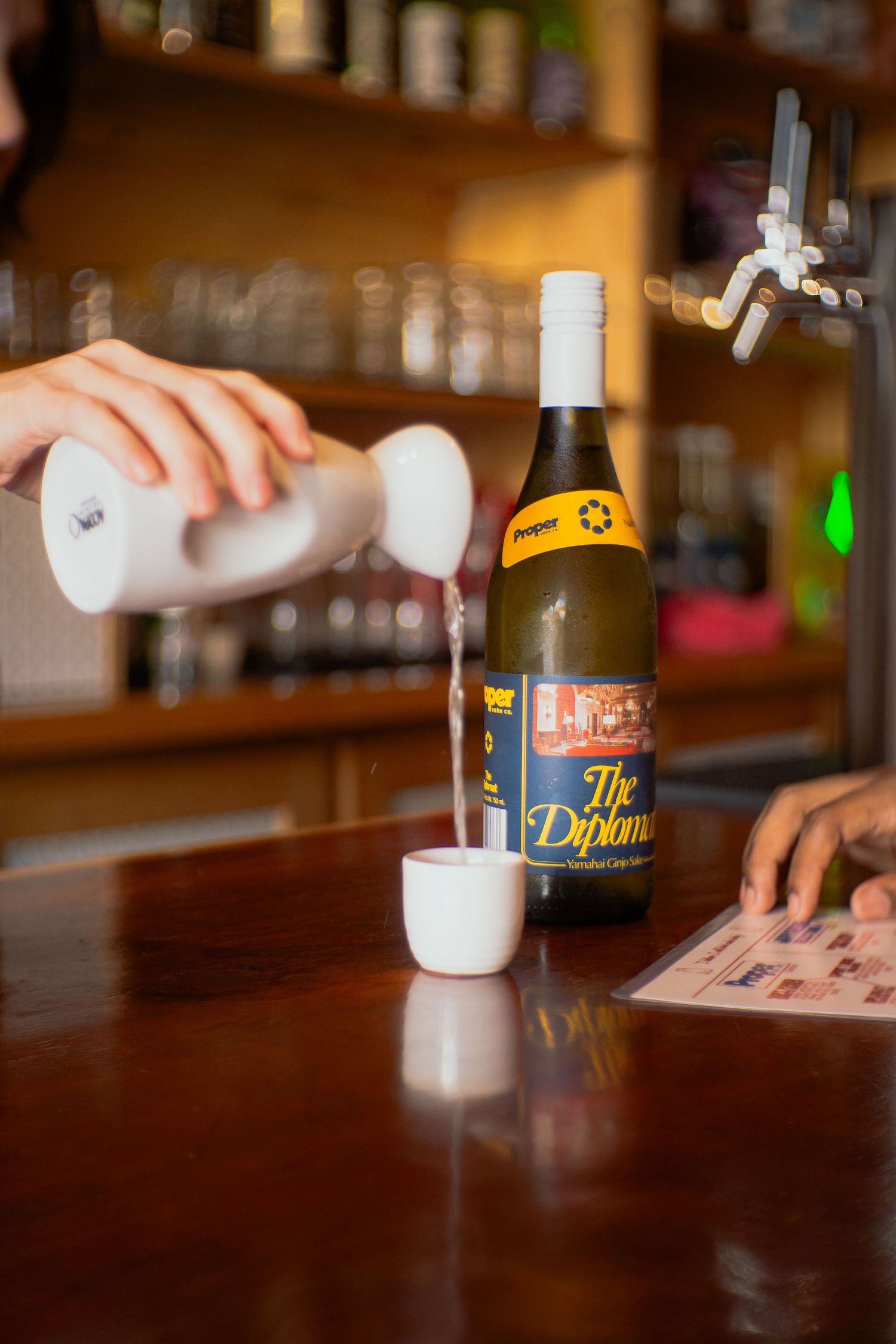 Four sequential shots of a beer bottle and glass being poured at a wooden bar counter in dim lighting.