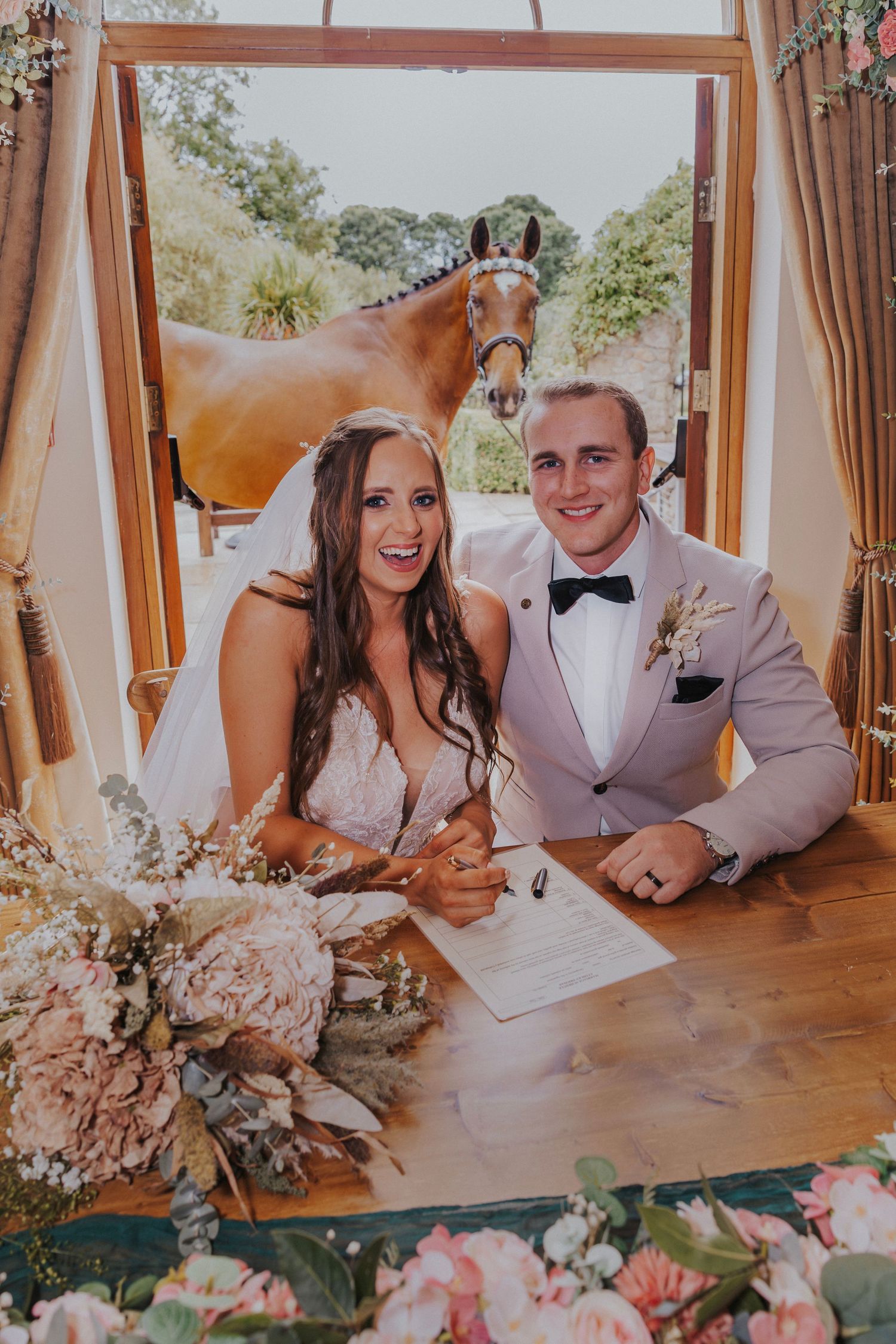 Couple signing marriage documents at wedding ceremony with pink floral decor and elegant indoor setting.