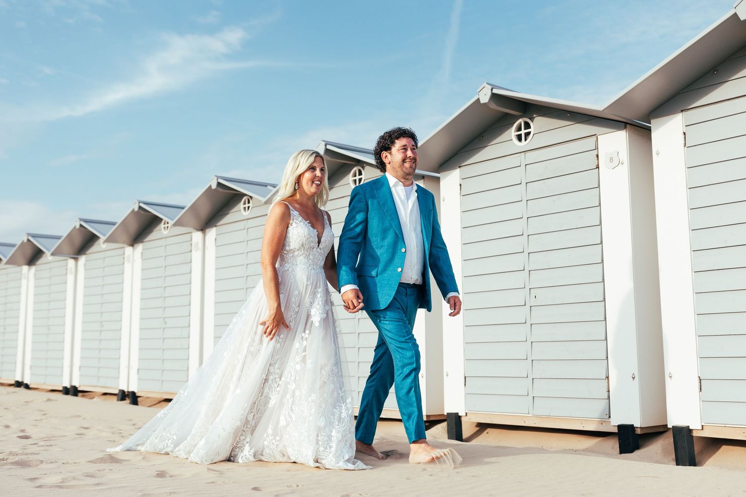 Bruid en bruidegom, op blote voeten in het zand. Lopend langs strandhuisjes.