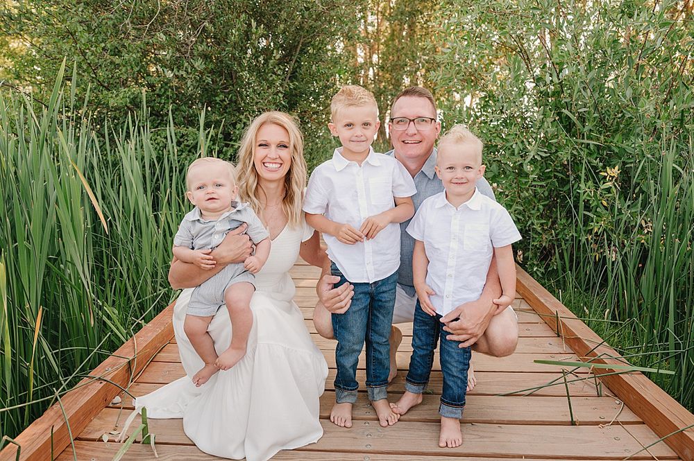 A family poses together on a wooden boardwalk surrounded by tall reeds and greenery on a sunny day.