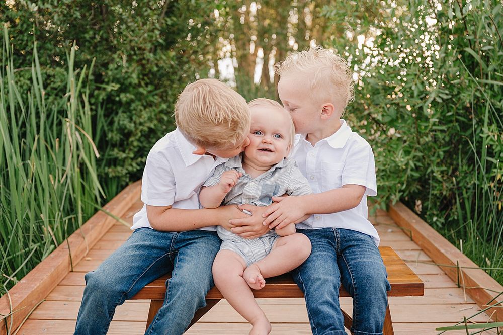Three children share sweet moments together while sitting on a wooden bridge surrounded by lush greenery.