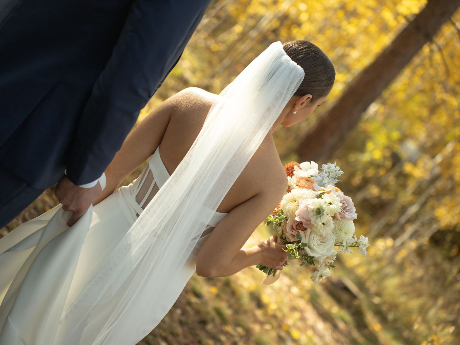 A bride in a white wedding dress holds a pastel bouquet against a backdrop of autumn foliage.
